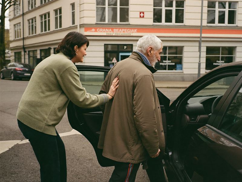 Betreuungskraft hilft einem älteren Herrn beim Aussteigen aus dem Auto vor einer Arztpraxis