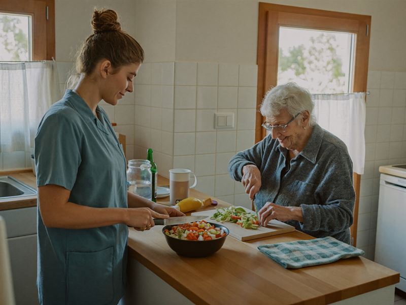 Betreuungskraft und ältere Frau schneiden gemeinsam Gemüse in einer hellen Küche